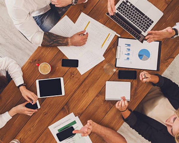 Group of busy business people working in office, top view of wooden table with mobile phones, laptop, tablet and documents papers with diagram. Men and women team have brainstorm discussion, vertical
